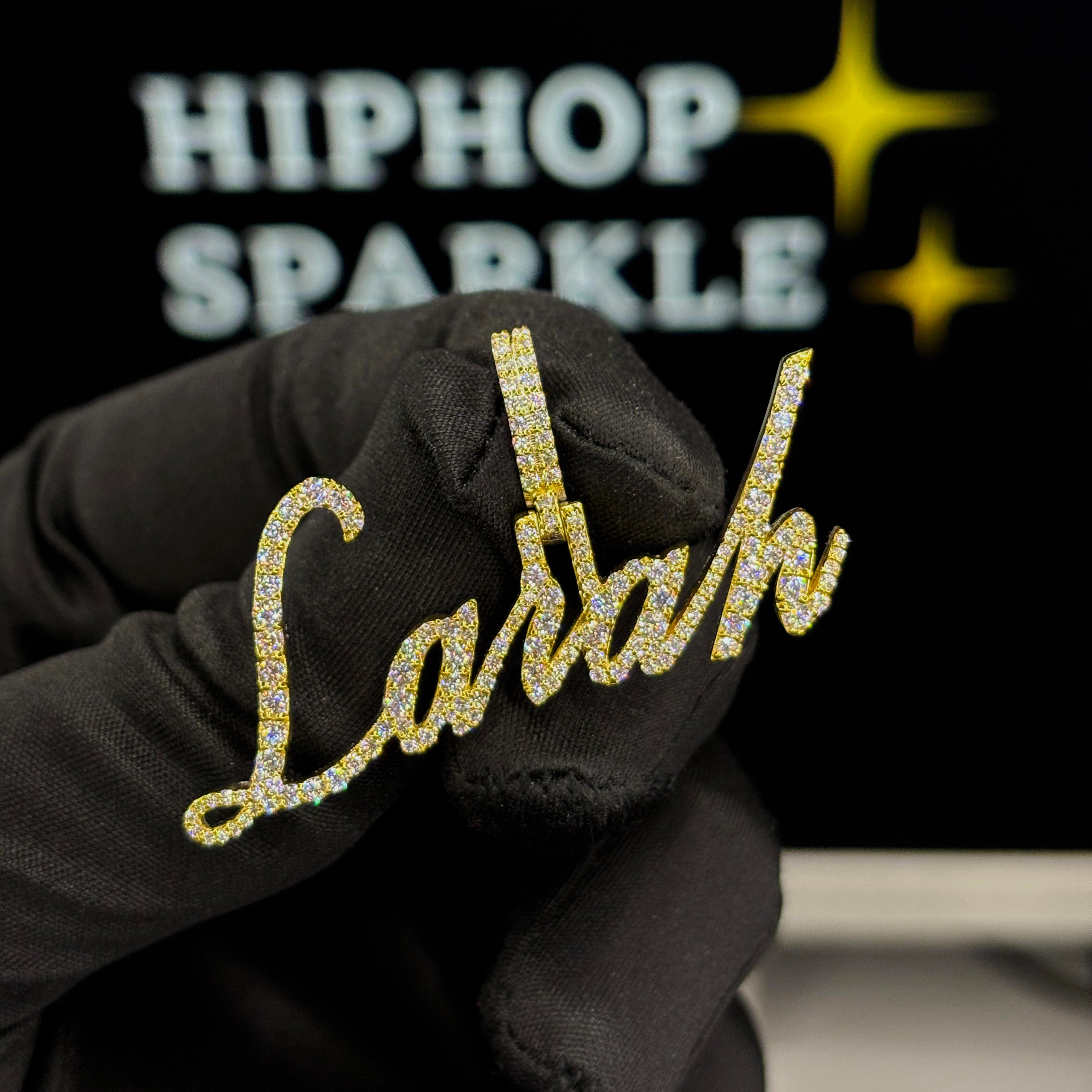 Iced-out gold script name pendant reading Carah, covered with moissanite stones and held with black gloves – Hip Hop Sparkle jewelry